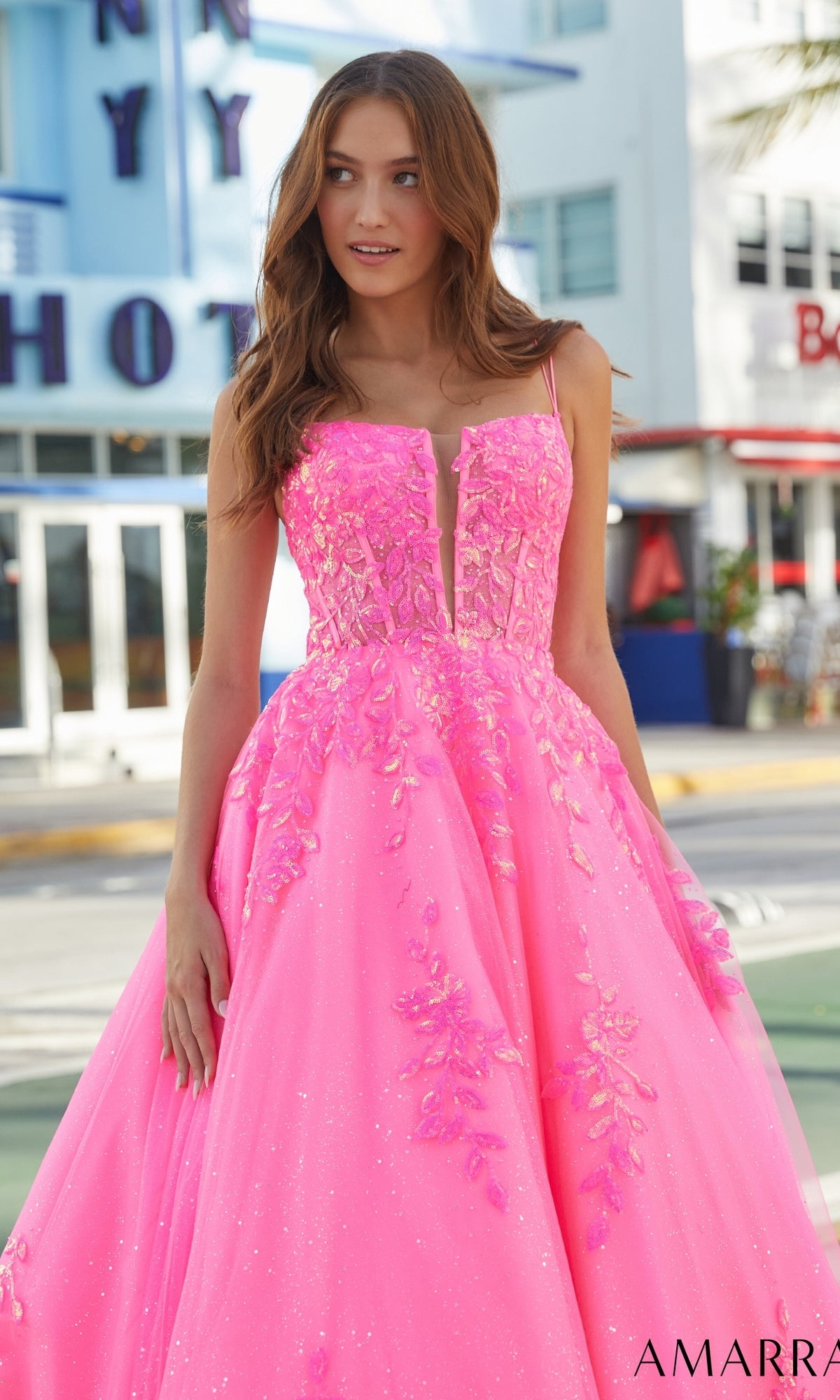 A woman wears the Amarra Long Prom Dress 88574, a bright pink gown with floral appliqué and corset bodice, standing outdoors in front of a building with